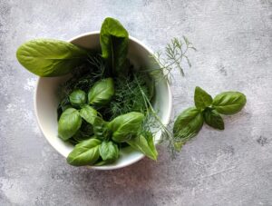 A variety of fresh green herbs including basil in a white bowl on a textured surface.