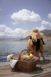 Woman enjoying a lakeside picnic with a basket of fruits and flowers.